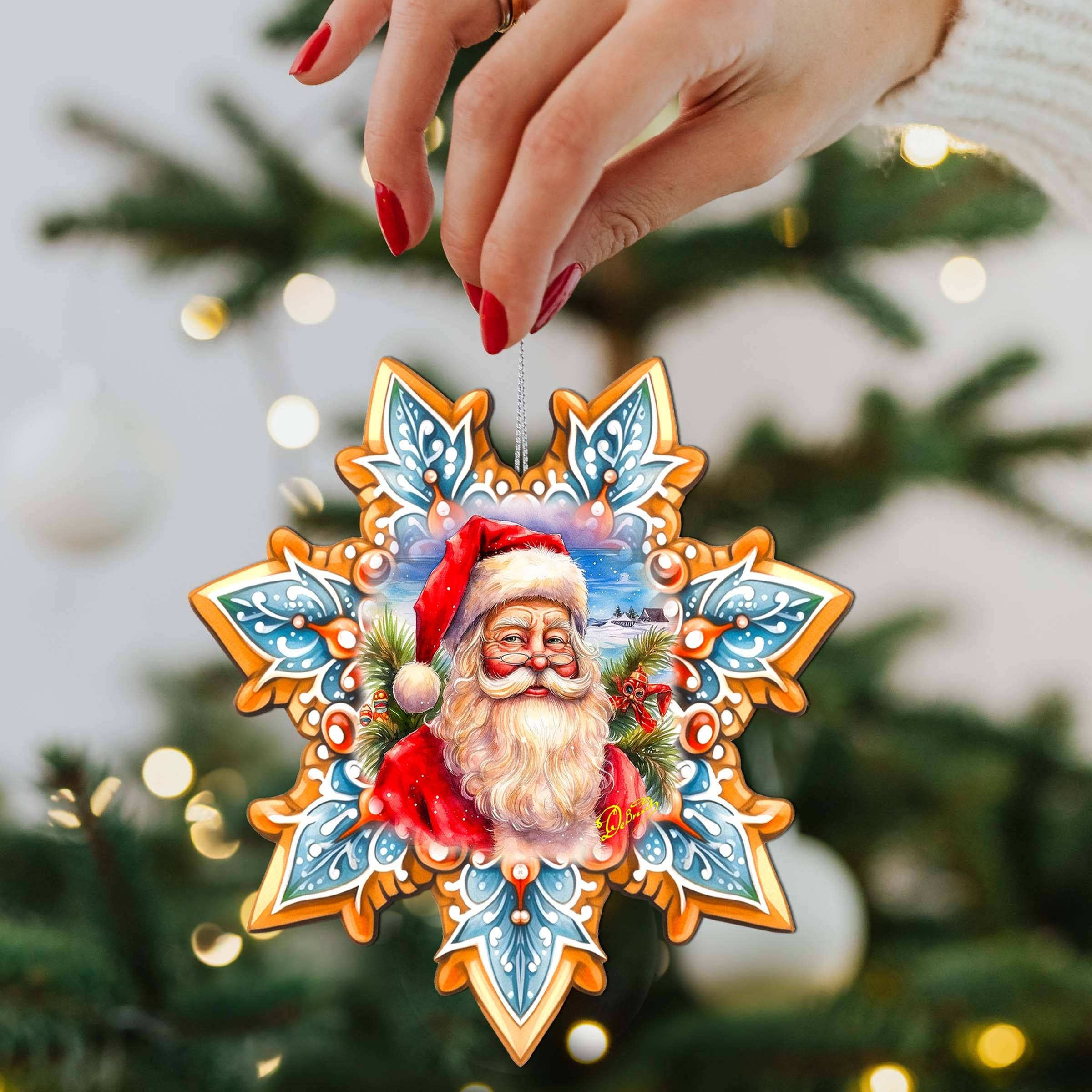 A hand holds an Ornament: Santa Snowflake Wood Ornaments GDebrekht in front of a beautifully lit and decorated Christmas tree.