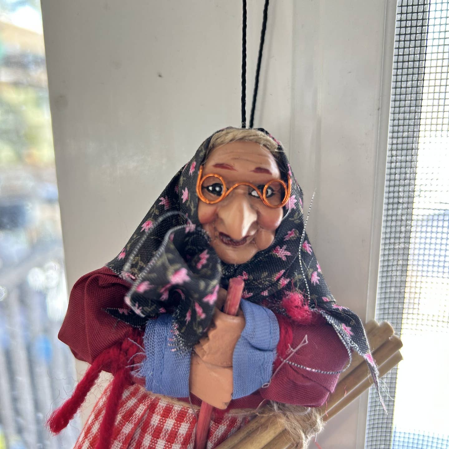 The Kitchen Witch Doll Opalina Red, 8.3" tall, features glasses, a headscarf, and a red checked apron. Hang this charming decor near your window for a traditional good luck touch in your kitchen.