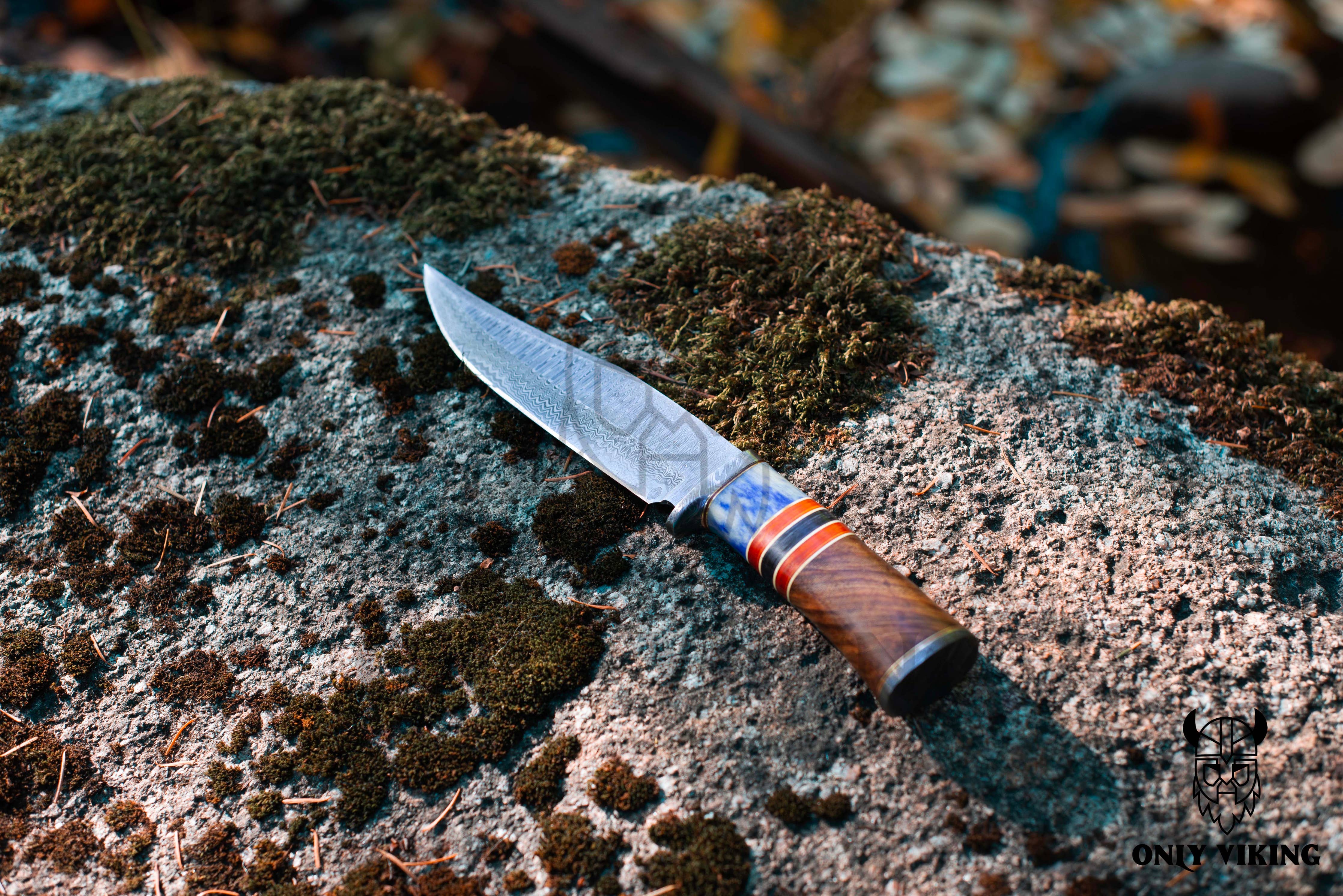 The Damascus Steel Hunting Knife, featuring a patterned blade and a decorative handle, rests on a mossy rock outdoors.