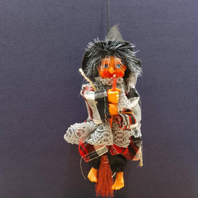 The Kitchen Witch: Minna Red Doll features a handmade orange face, hooded cloak, scarf, and holds a stick, displayed on a plain dark blue background—a charming piece of folklore décor.
