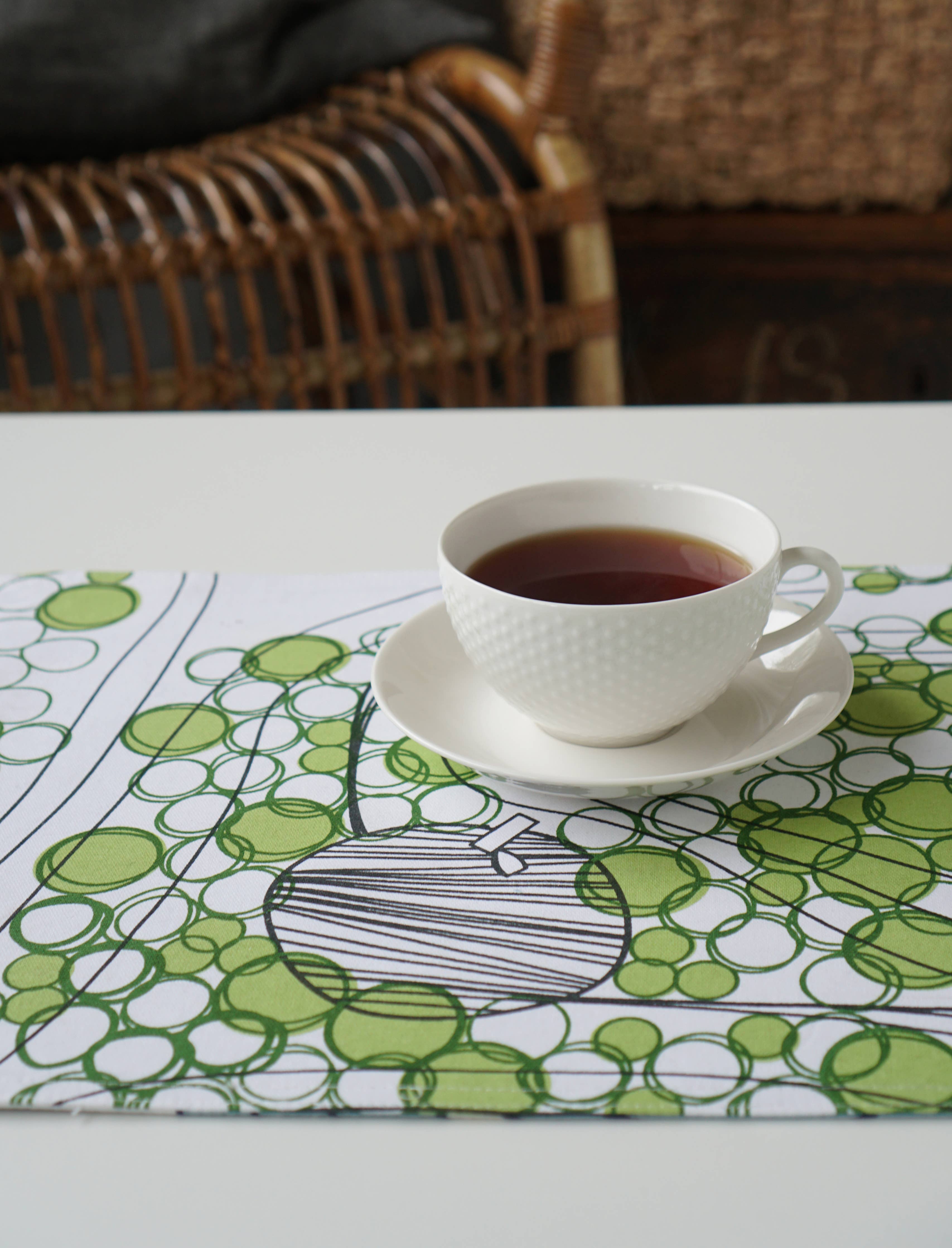 A cup of tea on a white saucer rests on the table, set against a double-sided placemat from Born in Sweden, featuring green and white patterns at 47cm x 34cm, beside a wicker chair.