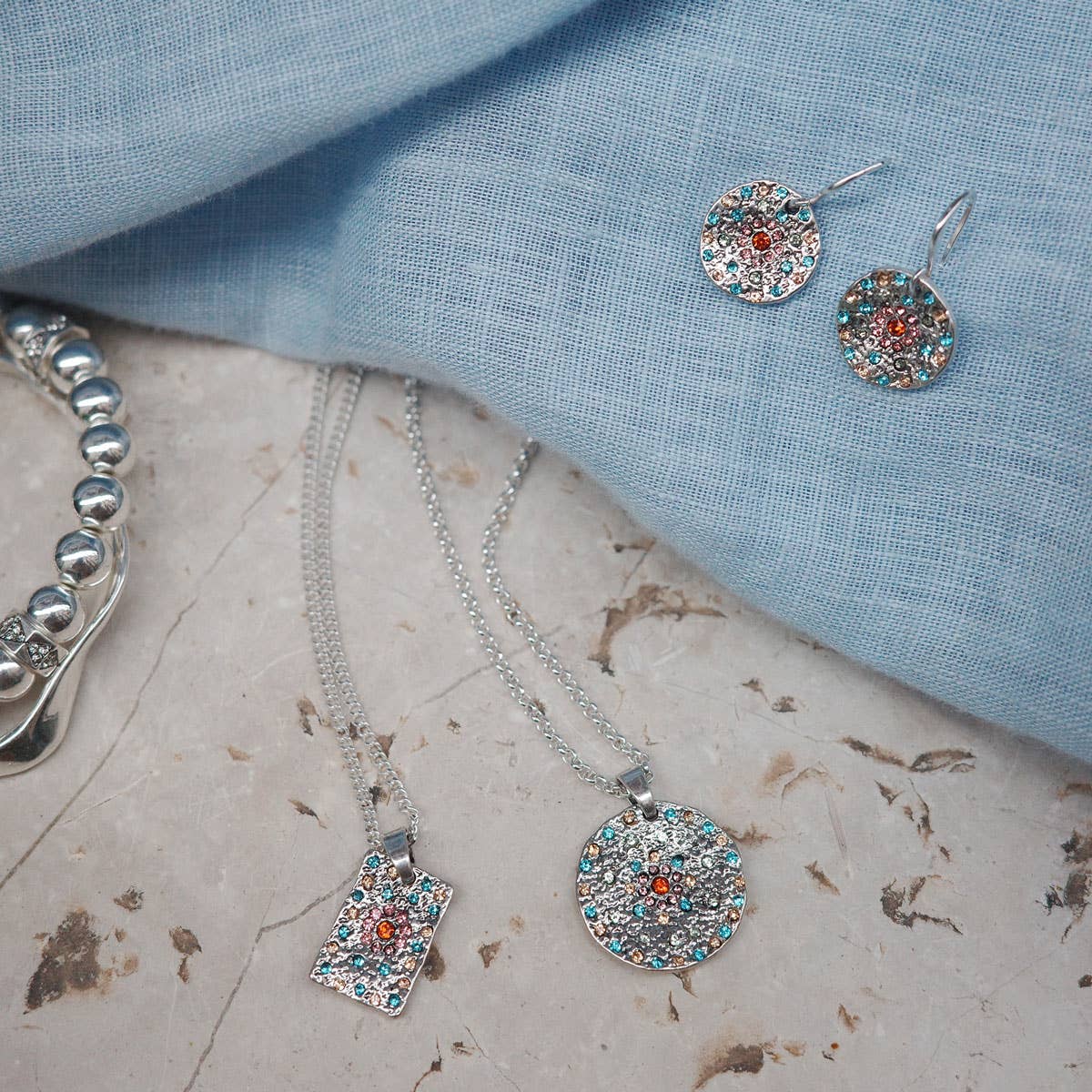 A&C Oslo Vibrance Silver Coin Necklace set, featuring a striking silver necklace, earrings, and a bracelet, rests elegantly on textured surfaces and blue fabric, radiating timeless elegance.