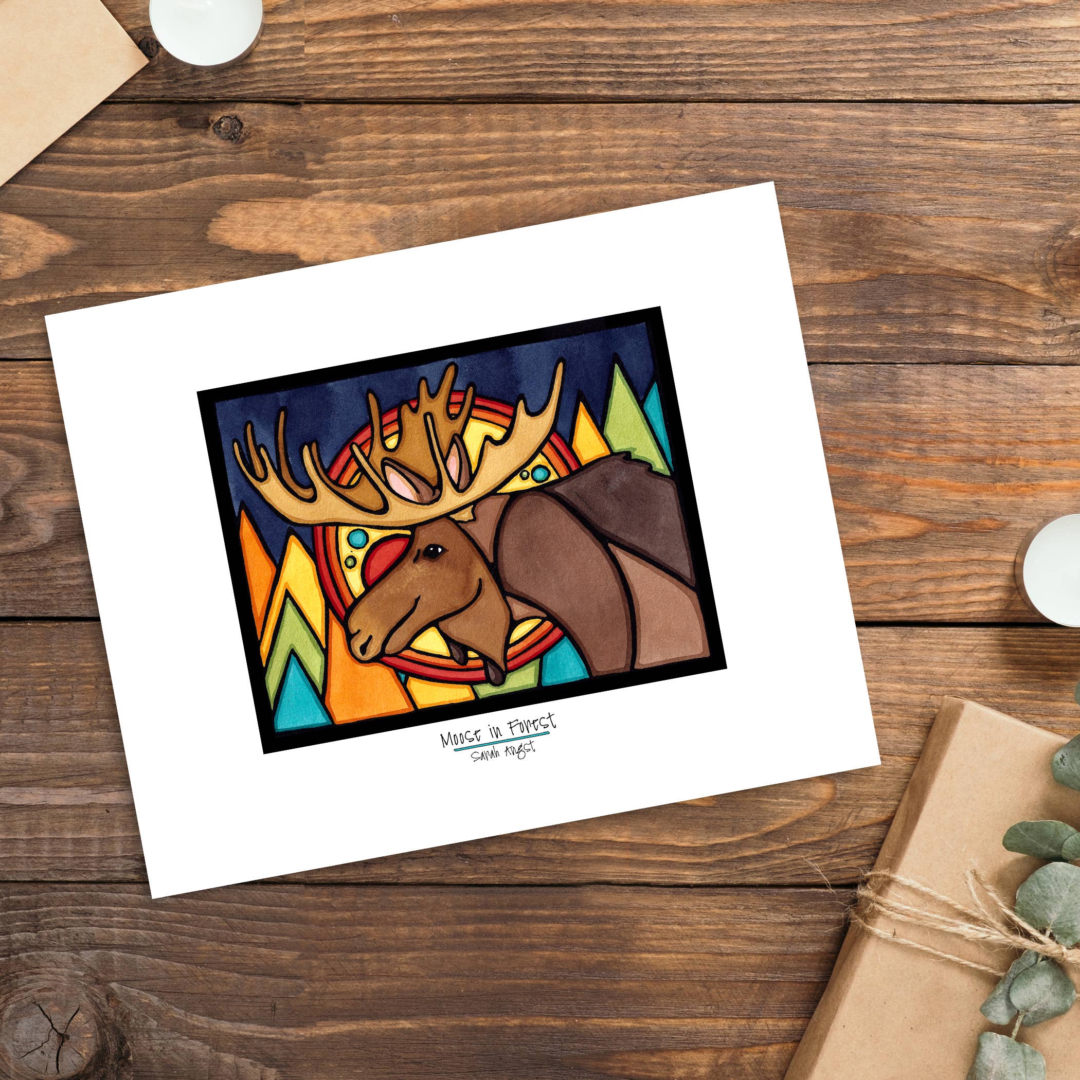 The 11"x14" "Moose in Forest" artwork by Sarah Angst, displaying stained glass-like colors, is framed and set on a wooden surface alongside a small plant and a wrapped gift.