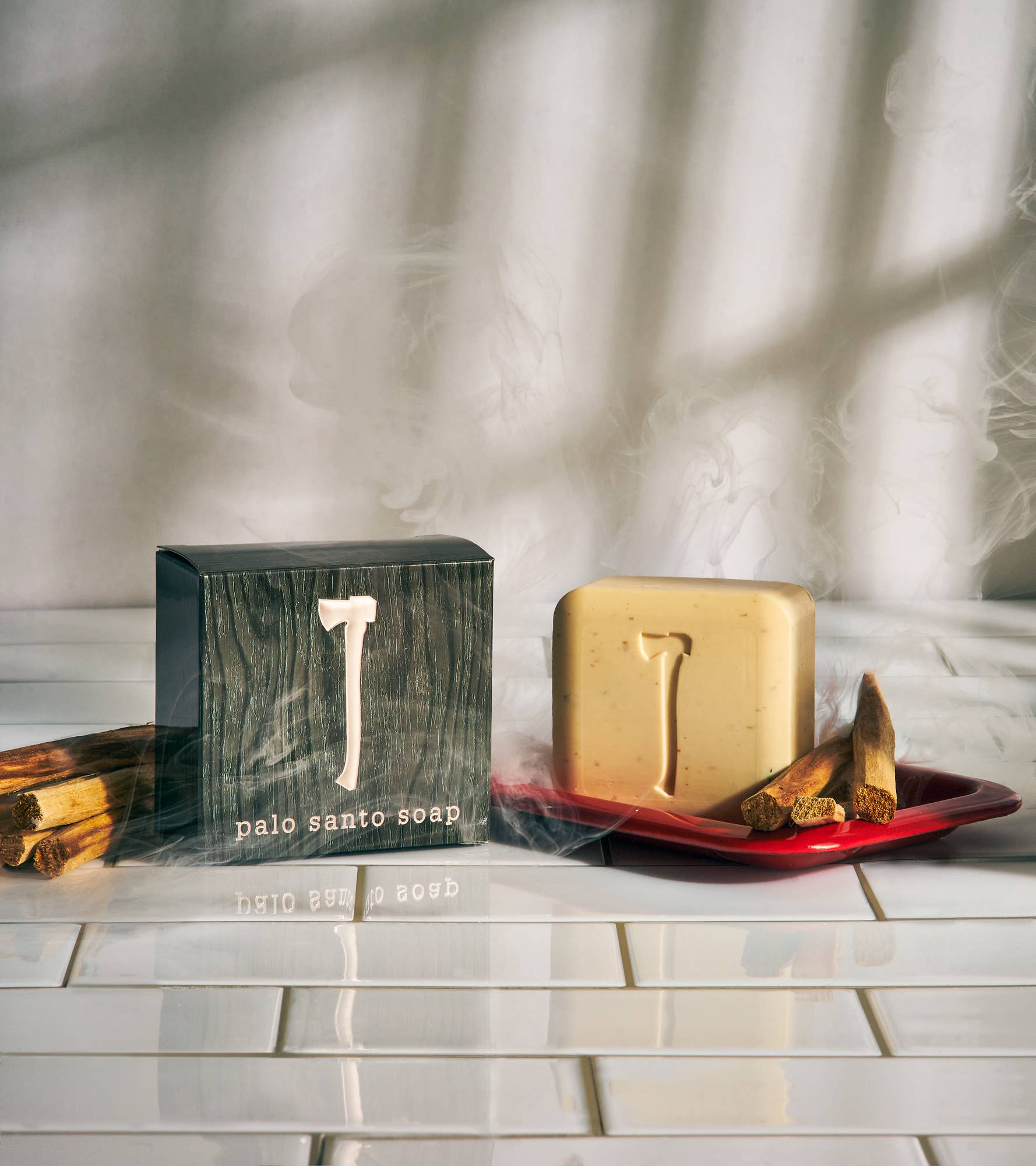 A box labeled "Soap: Palo Santo Soap" and an exfoliating bath bar featuring a raised design rest atop a red dish alongside palo santo sticks, enveloped in gentle smoke on a white tiled surface.