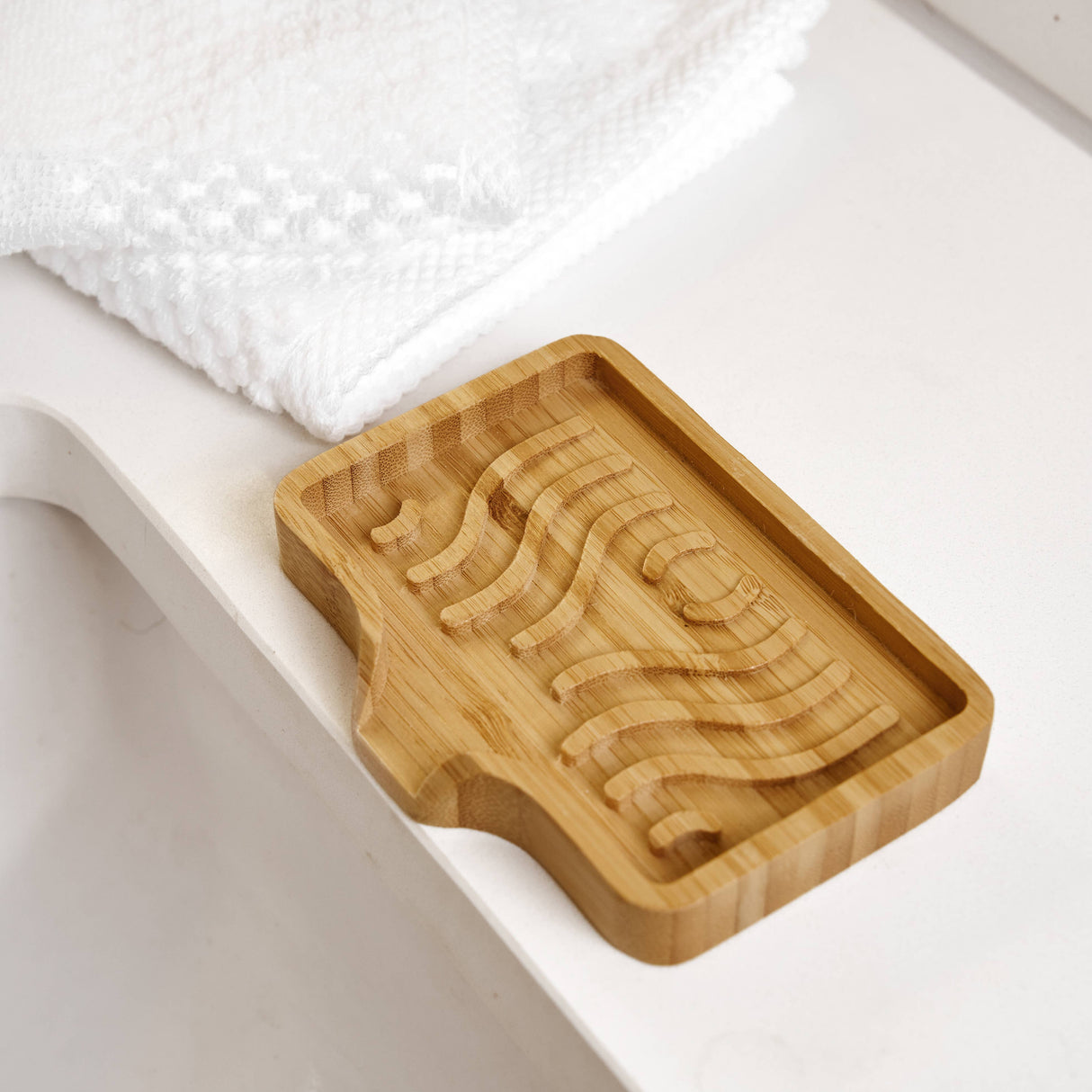 Bamboo soap lift with wavy grooves on a white bathroom countertop beside folded white towels.