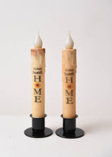 The Home Sweet Home Sunflower LED Timer Taper Candle: Top Dusted features two battery-operated tapers on black stands, each with a sunflower and phrase design, creating a cozy, charming ambiance against a white background.