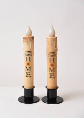 The Home Sweet Home Sunflower LED Timer Taper Candle: Top Dusted features two battery-operated tapers on black stands, each with a sunflower and phrase design, creating a cozy, charming ambiance against a white background.