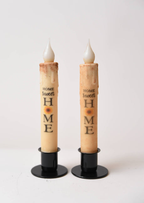 The Home Sweet Home Sunflower LED Timer Taper Candle: Top Dusted features two battery-operated tapers on black stands, each with a sunflower and phrase design, creating a cozy, charming ambiance against a white background.