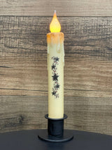 A battery-operated candle featuring a melted wax look and star decorations is displayed in a Round Wrought Iron Taper Candle Holder, set against a wooden background.