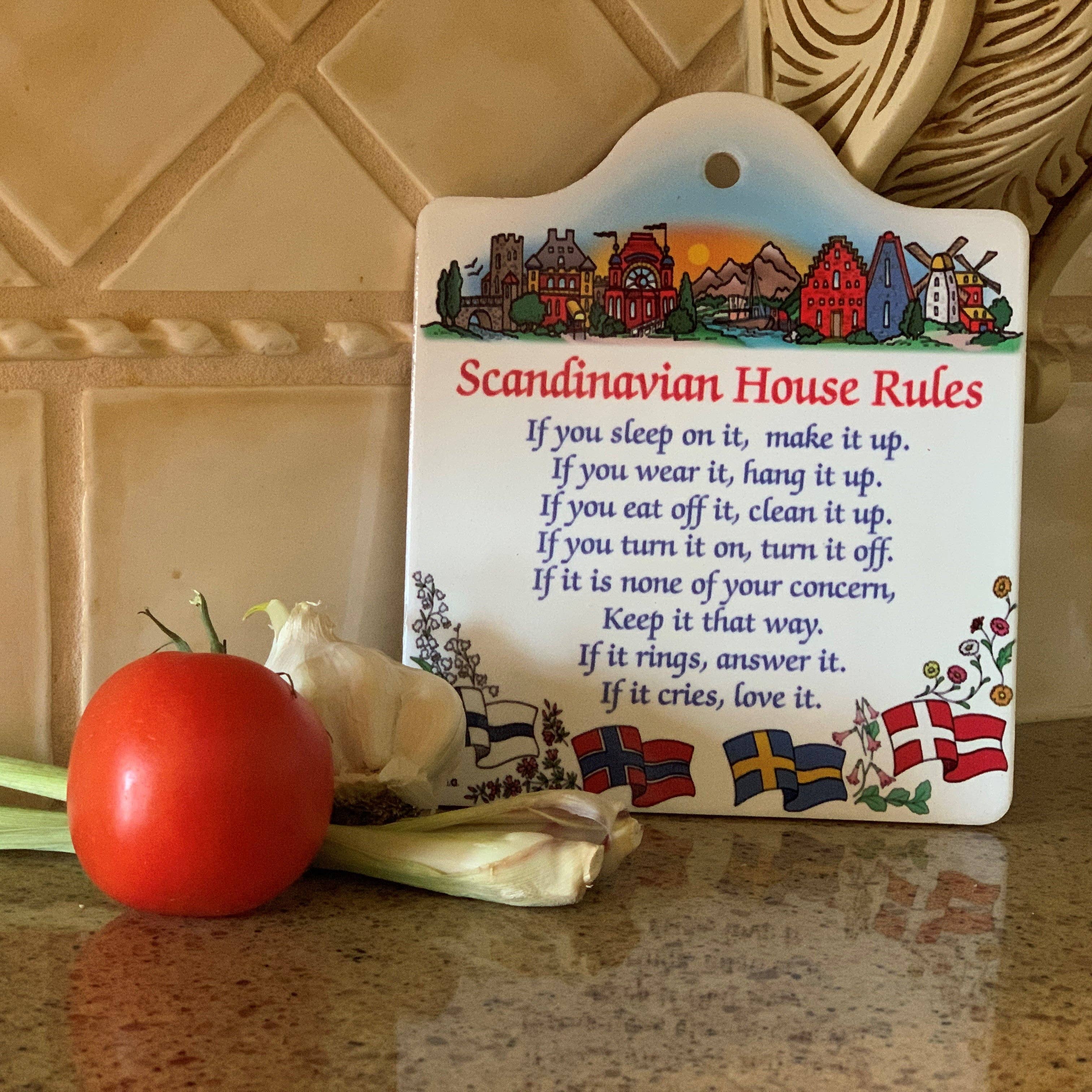 The Scandinavian House Rules decorative plaque, humorously detailing chores, is featured on the cork-backed ceramic cheeseboard titled Tile, nestled between a tomato and garlic. Flags depicted at the bottom add a playful element to your kitchen decor.