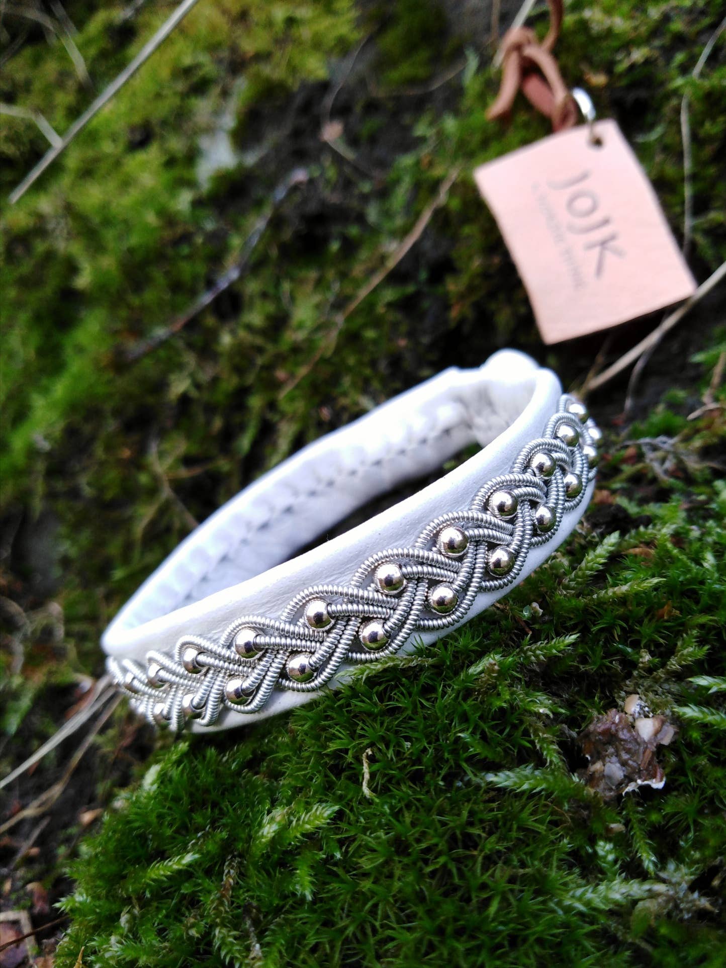 A Sami Lapland bracelet with pewter, silver, and gold beads on black reindeer hide (17-18 cm) is displayed on green moss with a brown Joik tag.