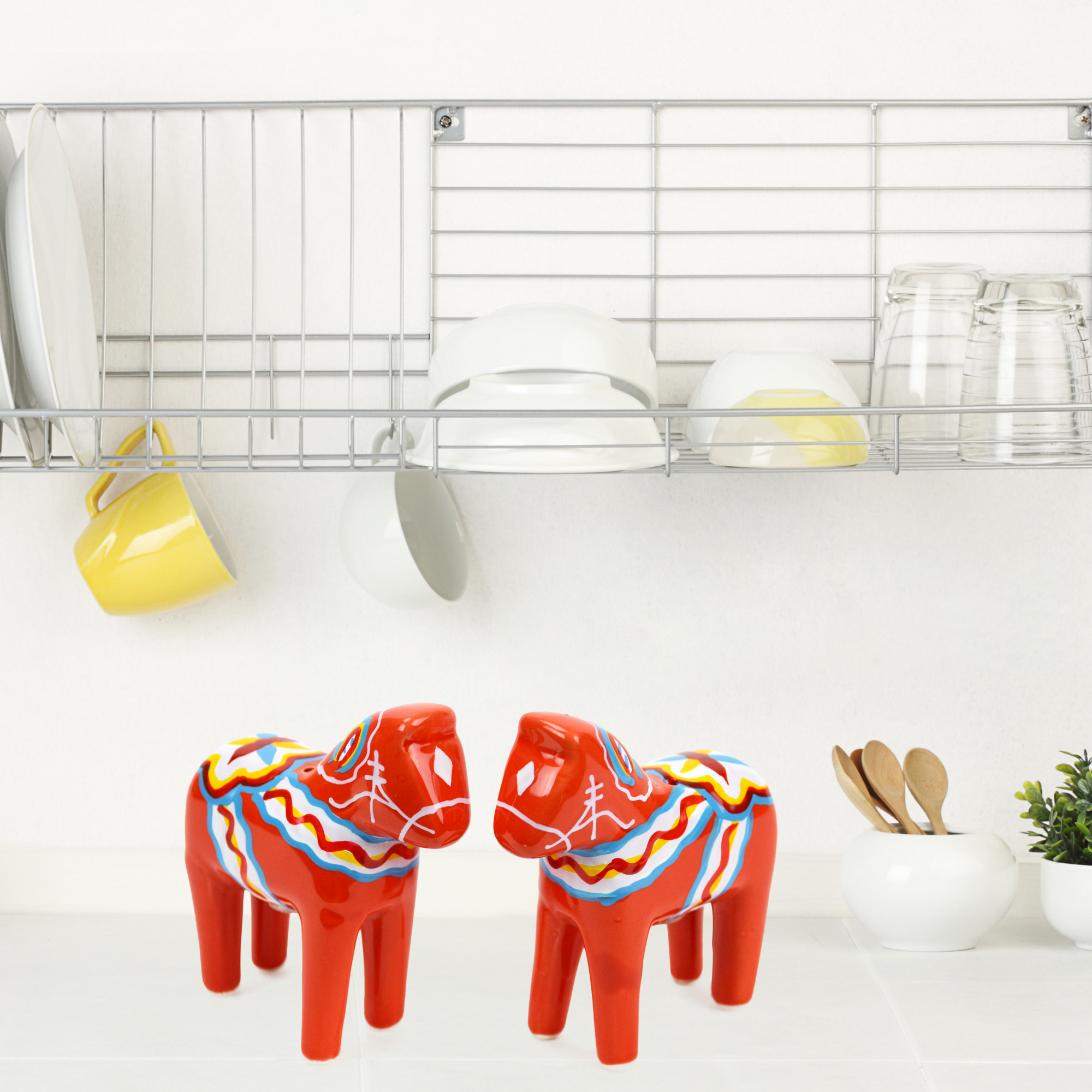 Two red Shakers: Dala Horse Salt and Pepper shakers embody Swedish charm atop a colorful countertop. Above, a dish rack holds white and yellow dishes, cups, and glasses. On the right, wooden spoons in a container add to the scenes Scandinavian heritage.