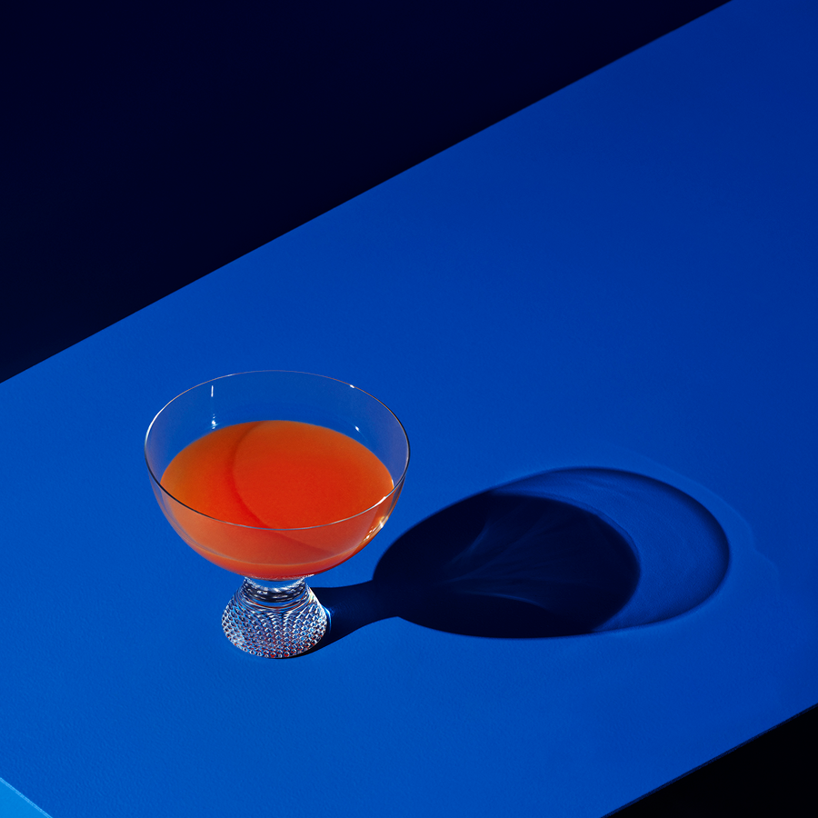 The KostaBoda: Limelight Bowl On Foot, filled with orange liquid, casts a striking shadow on a vivid blue surface in dramatic lighting.