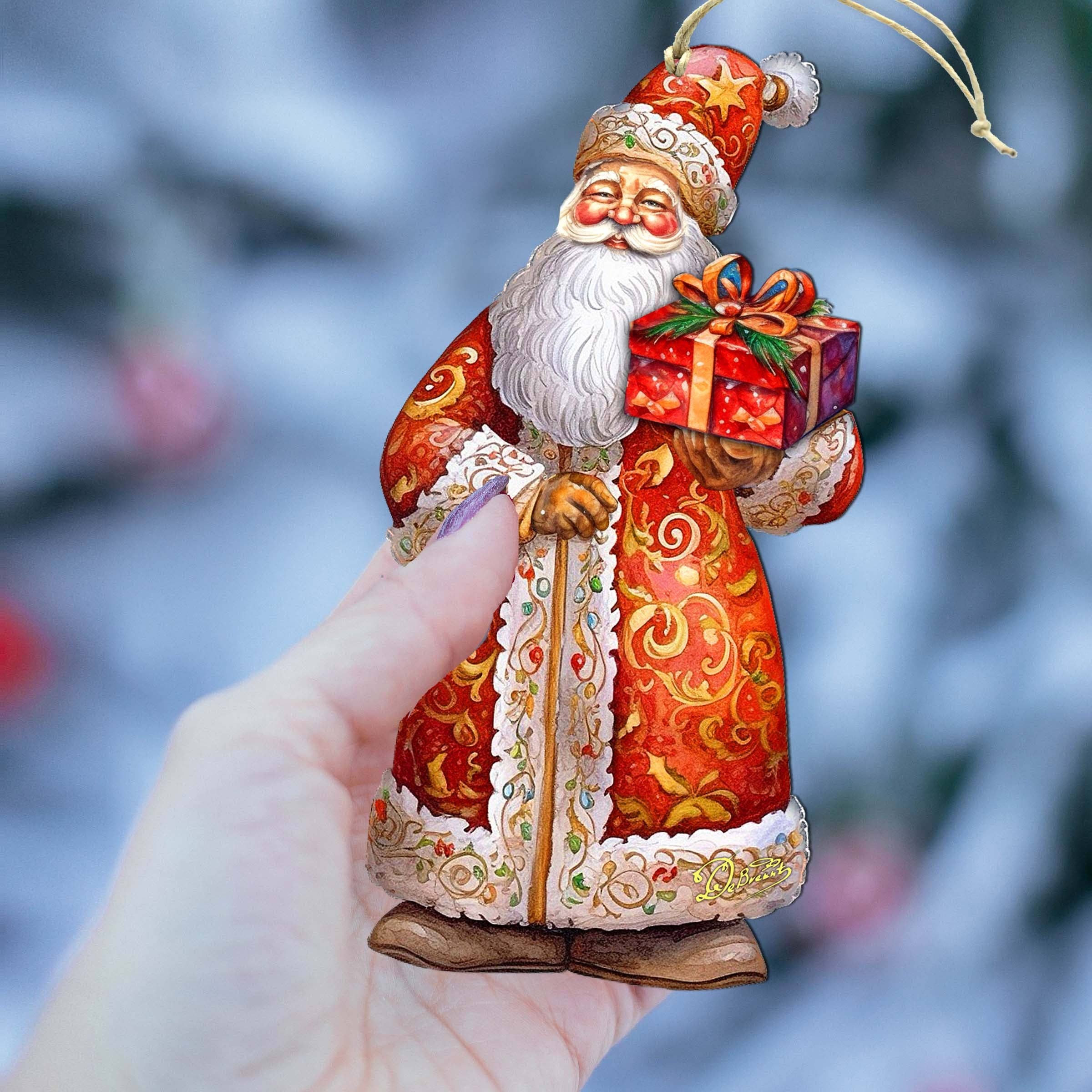 A hand holds an ornament of a figure in a red robe with a gift box, capturing the essence of the Ornament: St. Nick's Gift Wood Ornaments GDebrekht Christmas Santa against a blurred snowy background, perfect for your Christmas Holiday Decor.