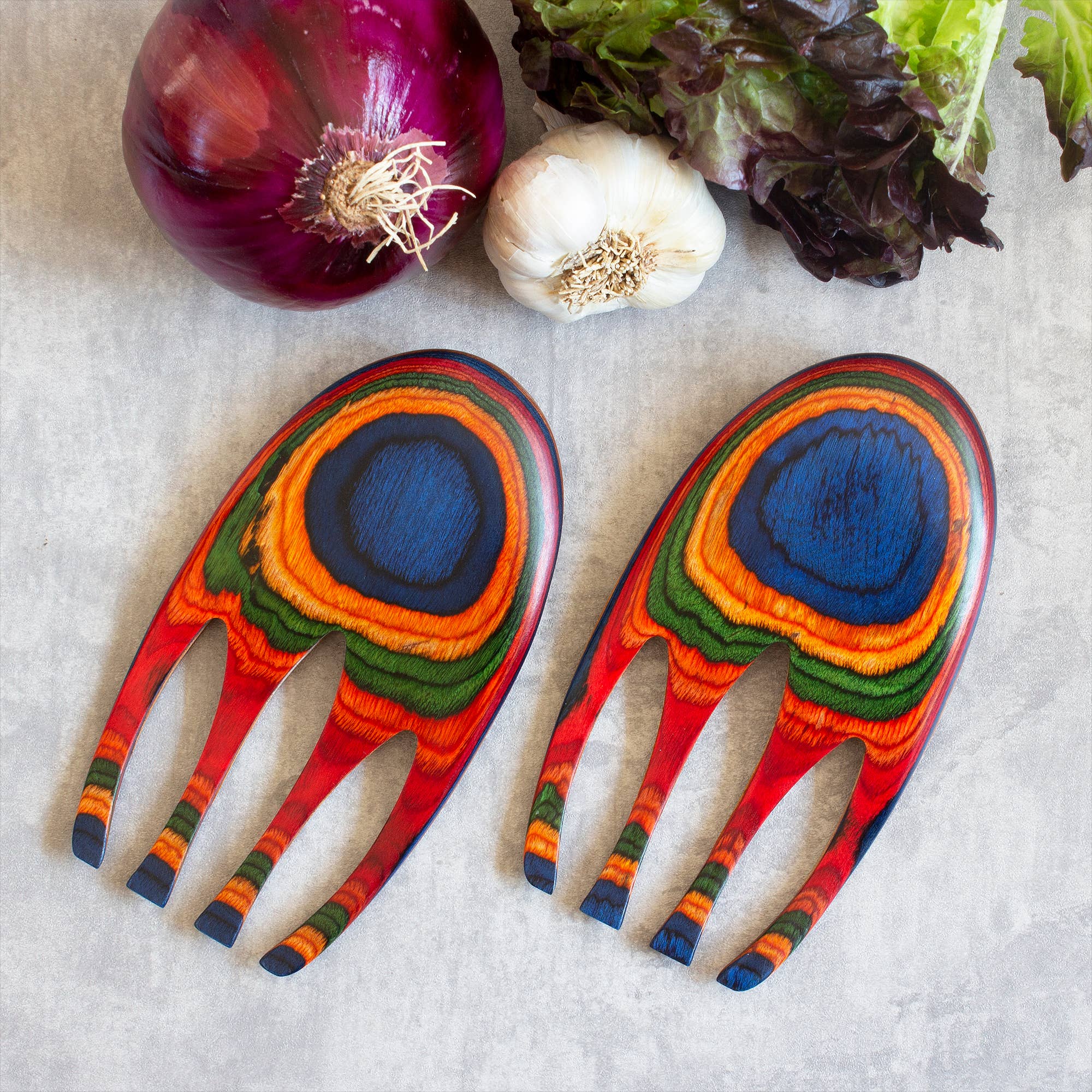 The Marrakesh Salad Hands, a set of multicolored wooden salad serving utensils, rest on a gray surface alongside a red onion, a garlic bulb, and vibrant green leafy lettuce—ideal for effortlessly tossing pasta salads.