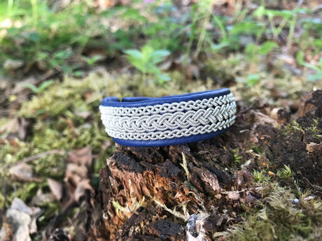 A black reindeer leather Sami bracelet (#5B) with pewter and silver thread sits on a mossy tree stump, green foliage in the background.