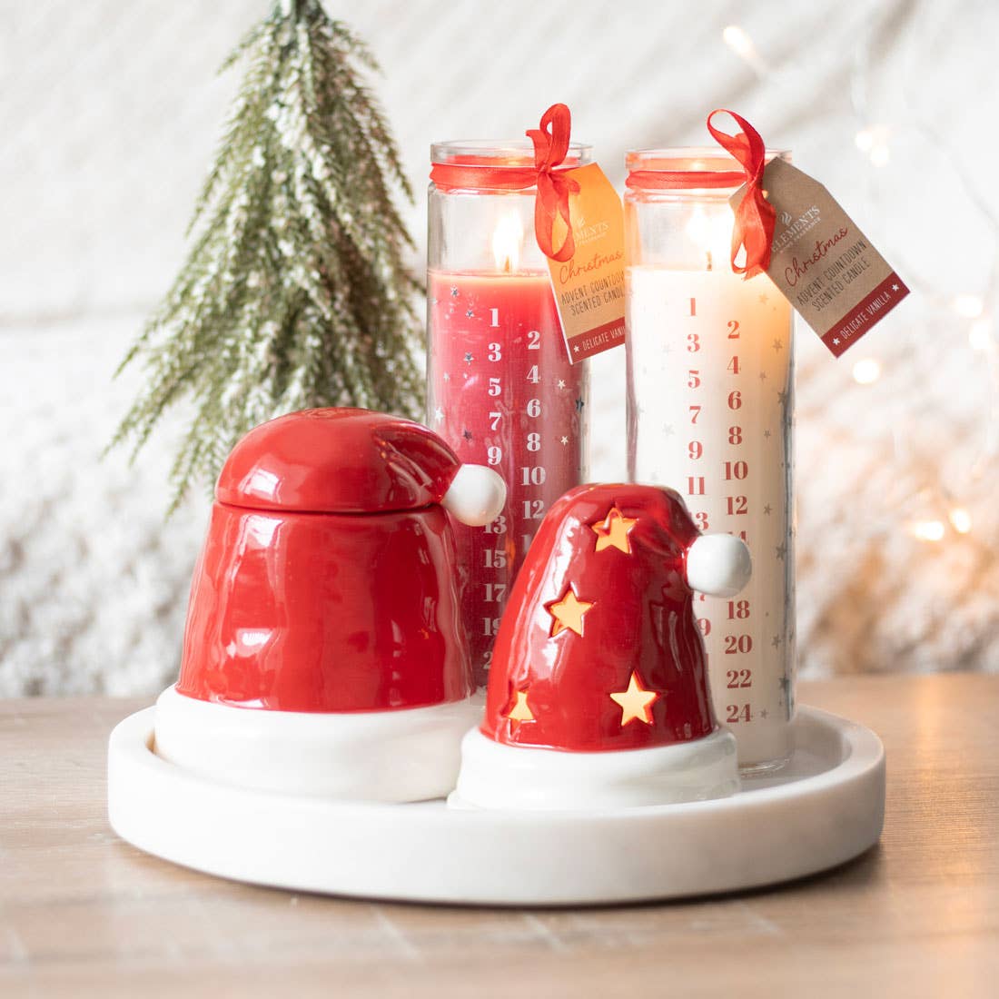 A display of three Red Vanilla Christmas Advent Tube Candles made from vegan paraffin wax in a color scheme of red and white is accompanied by two red Santa hat-shaped containers on a tray. In the background, a small tree enhances the cozy atmosphere with its subtle vanilla scent.