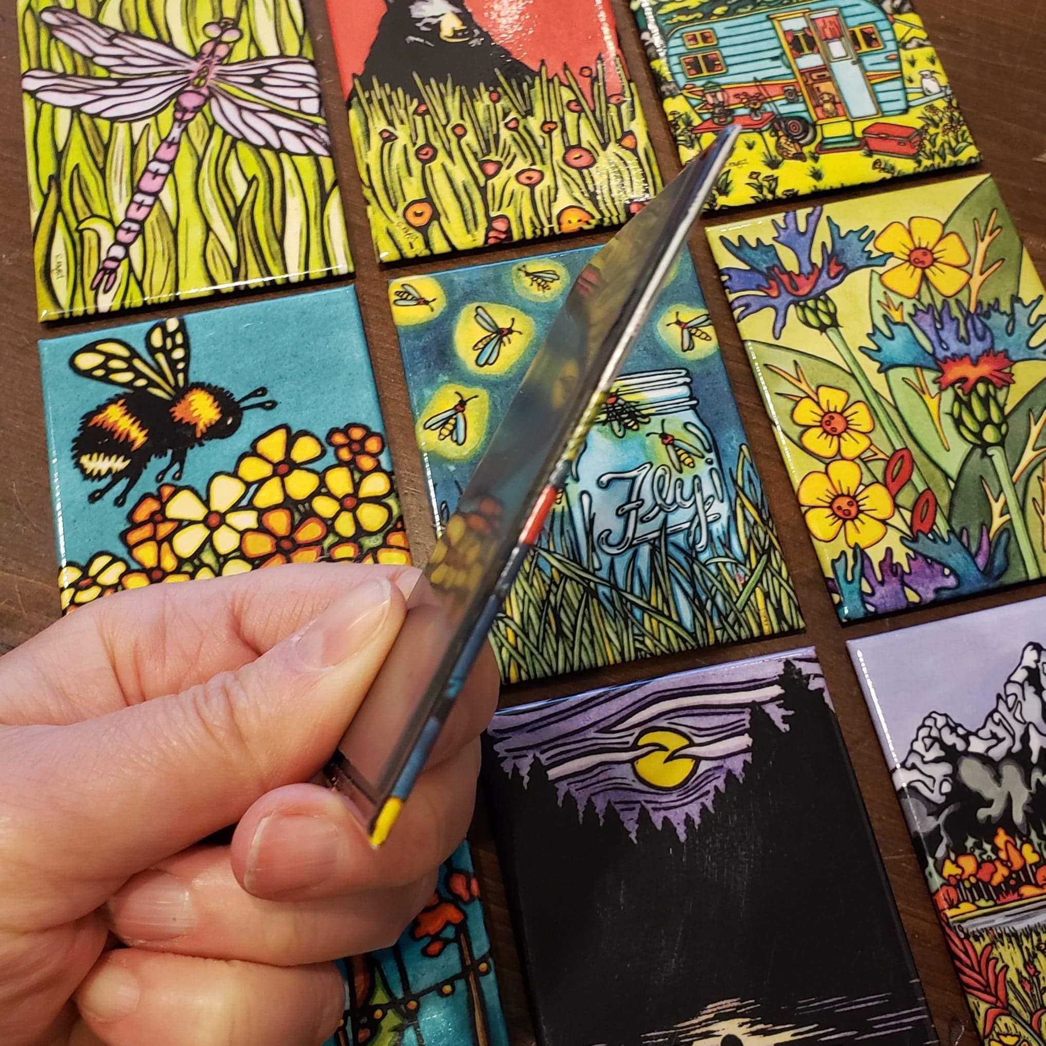 A hand displays a small card among colorful cards with the Minnesota Solitude Canoe Magnet and nature-themed illustrations, showcasing landscapes and insects, all artfully arranged on a table.