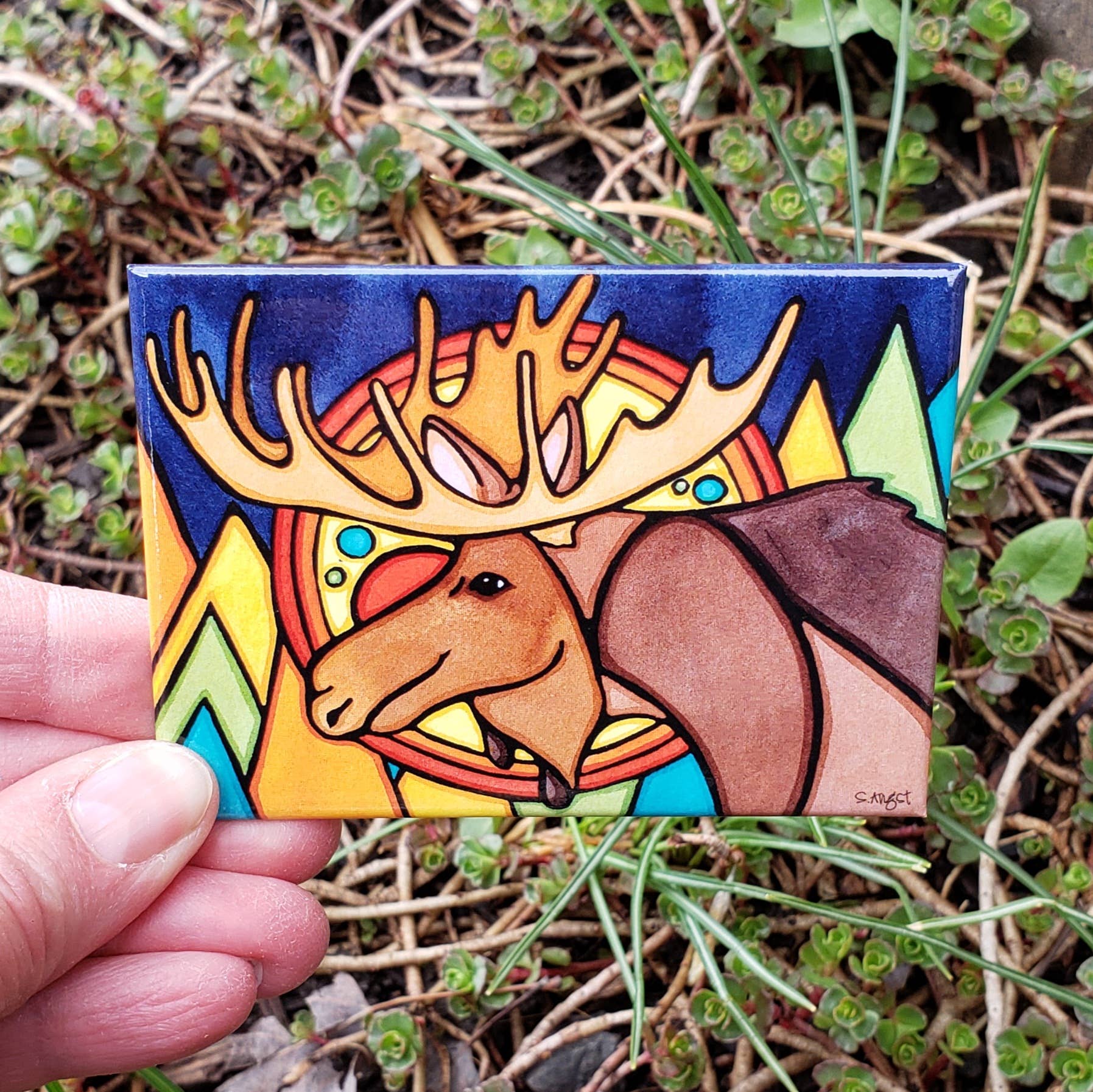 A hand holds the Forest Moose Magnet, featuring a vibrant painted moose with yellow antlers against a geometric backdrop of green triangles and a red-orange circle.