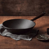 A Skeppshult Wok With Walnut Handle 12.2 / 30 cm sits on a cloth atop a wooden table, beside a plate of herbs and a small cup.