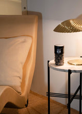 Beige chair with cushion beside marble-topped side table holding gold lamp, salty licorice jar, and capsules. Soft lighting, neutral interior.