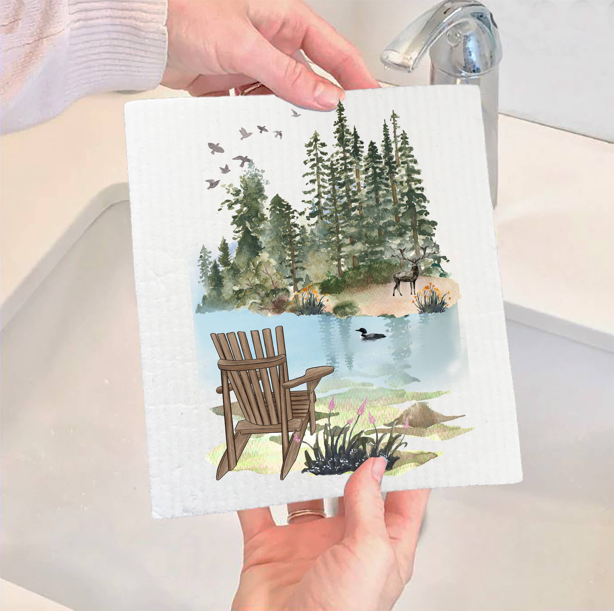 Hands holding a Swedish Dish Cloth: Andirondacks Chairs Lake Mountains, showcasing a chair by a lake with trees, birds, and a deer in a bathroom. This eco-friendly product blends elegant design with functionality.