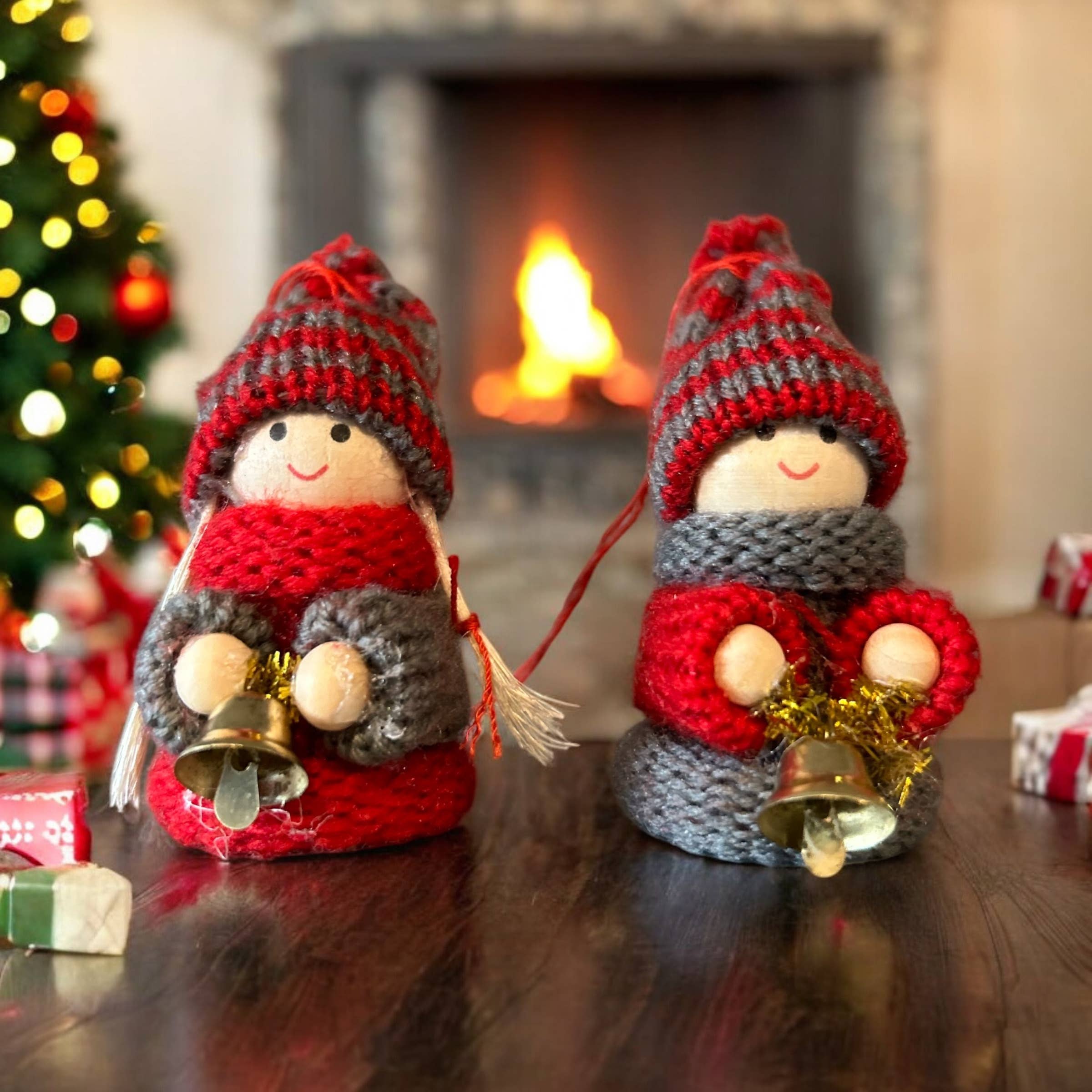 Two Tomte Children with Bells Christmas Tree Ornaments, wearing hats and scarves, sit on a table by the fireplace. The festive scene, complete with a decorated tree and gifts, makes these knit gnomes perfect for cozy holiday table decor.
