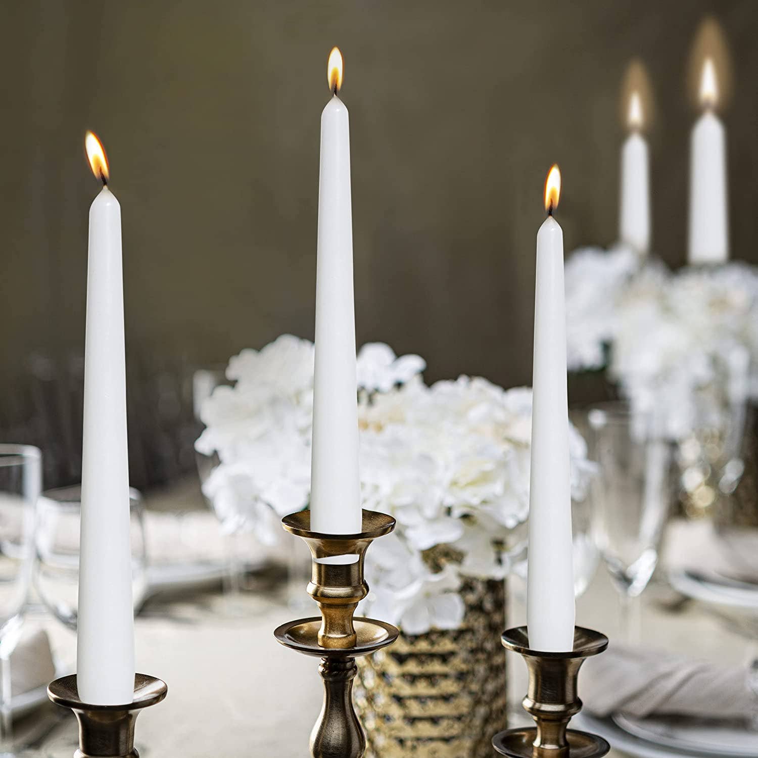 Three tall, unscented white taper candles from a 4-pack set glow softly on a brass candelabra, crafted from high-quality paraffin wax, against a blurred background featuring white flowers and elegant tableware.