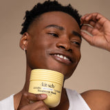 A person in a white tank top smiles and looks off to the side while holding a yellow container of Kitsch Hair: Moisturizing Coconut Oil Deep Conditioning Treatment Mask 10 oz, ready to enjoy its hydrating benefits.