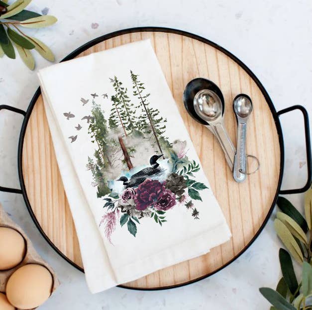 A round wooden tray features a charming Two Loons on a Lake Surrounded by Nature cotton tea towel, measuring spoons, and brown eggs. Perfect as a housewarming gift, plant leaves elegantly frame the scene.