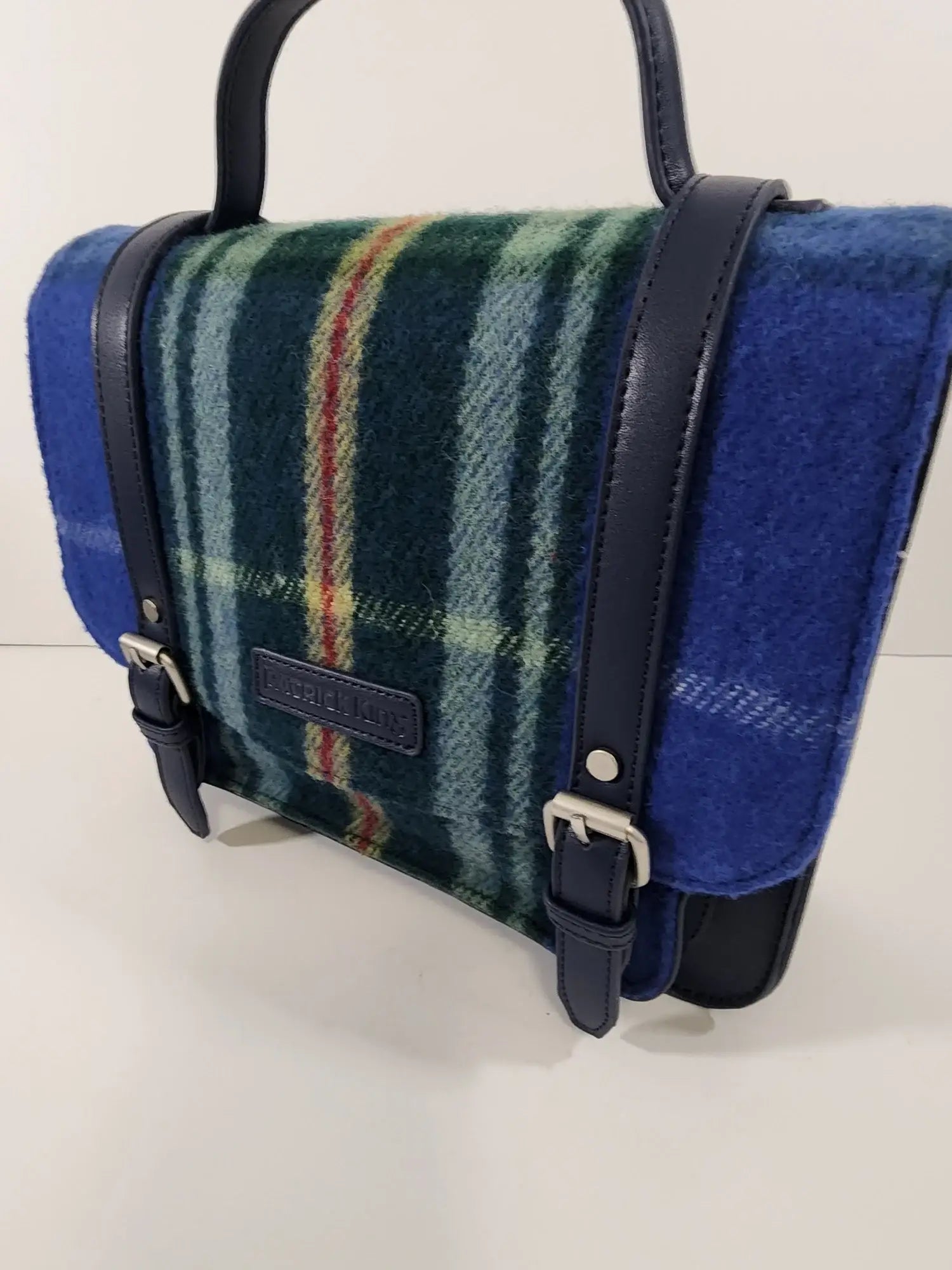 The Nova Scotia Patsy Handbag features blue and green plaid wool, dark blue leather straps, and silver buckles, displayed on a plain light background.