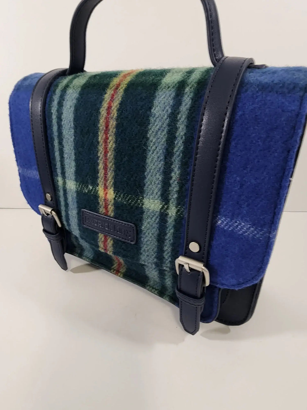 The Nova Scotia Patsy Handbag features blue and green plaid wool, dark blue leather straps, and silver buckles, displayed on a plain light background.