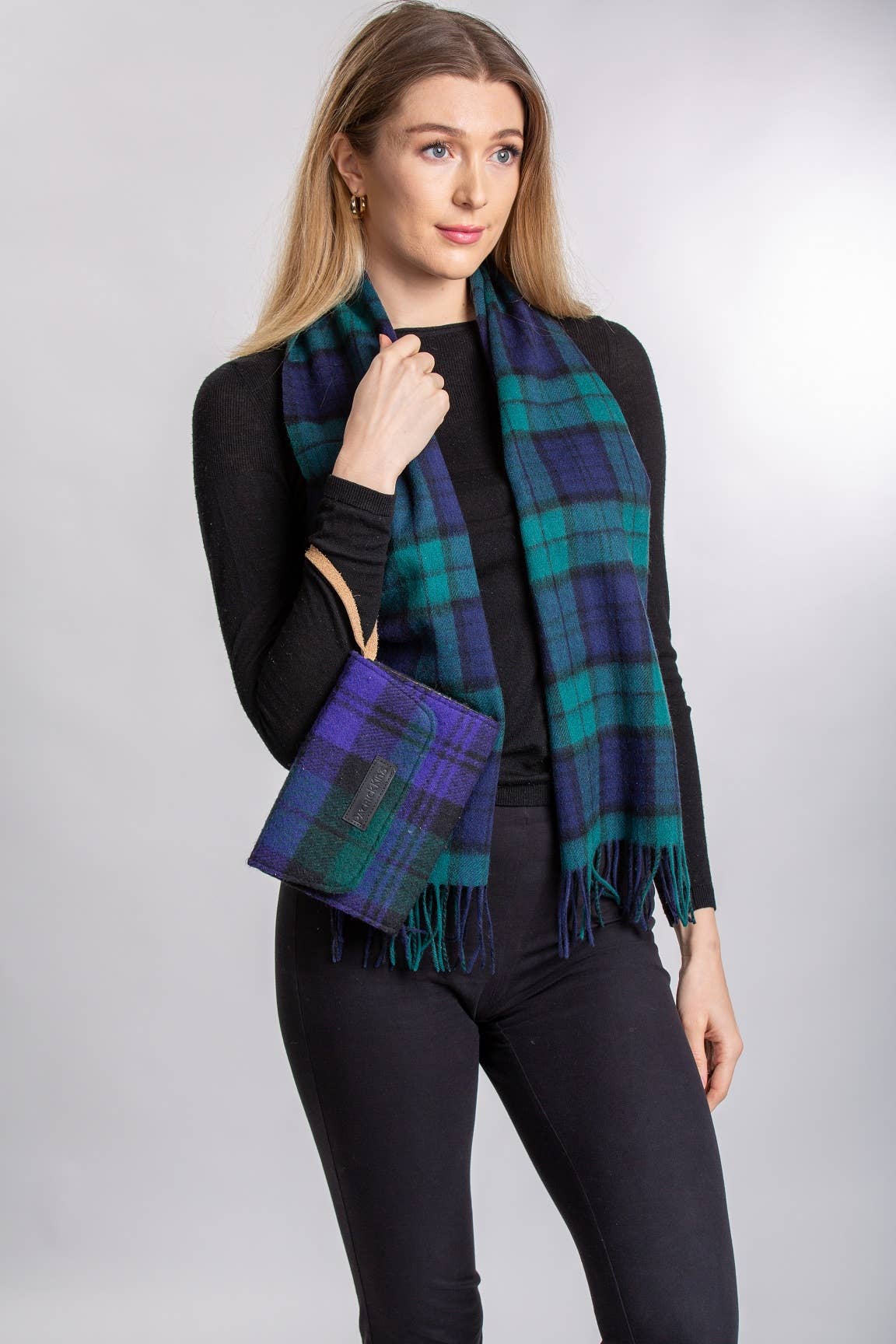 Woman in a black top and pants styles her look with a green and blue Blackwatch Lambswool Scarf by Patrick King, holding a matching plaid clutch against a plain background.