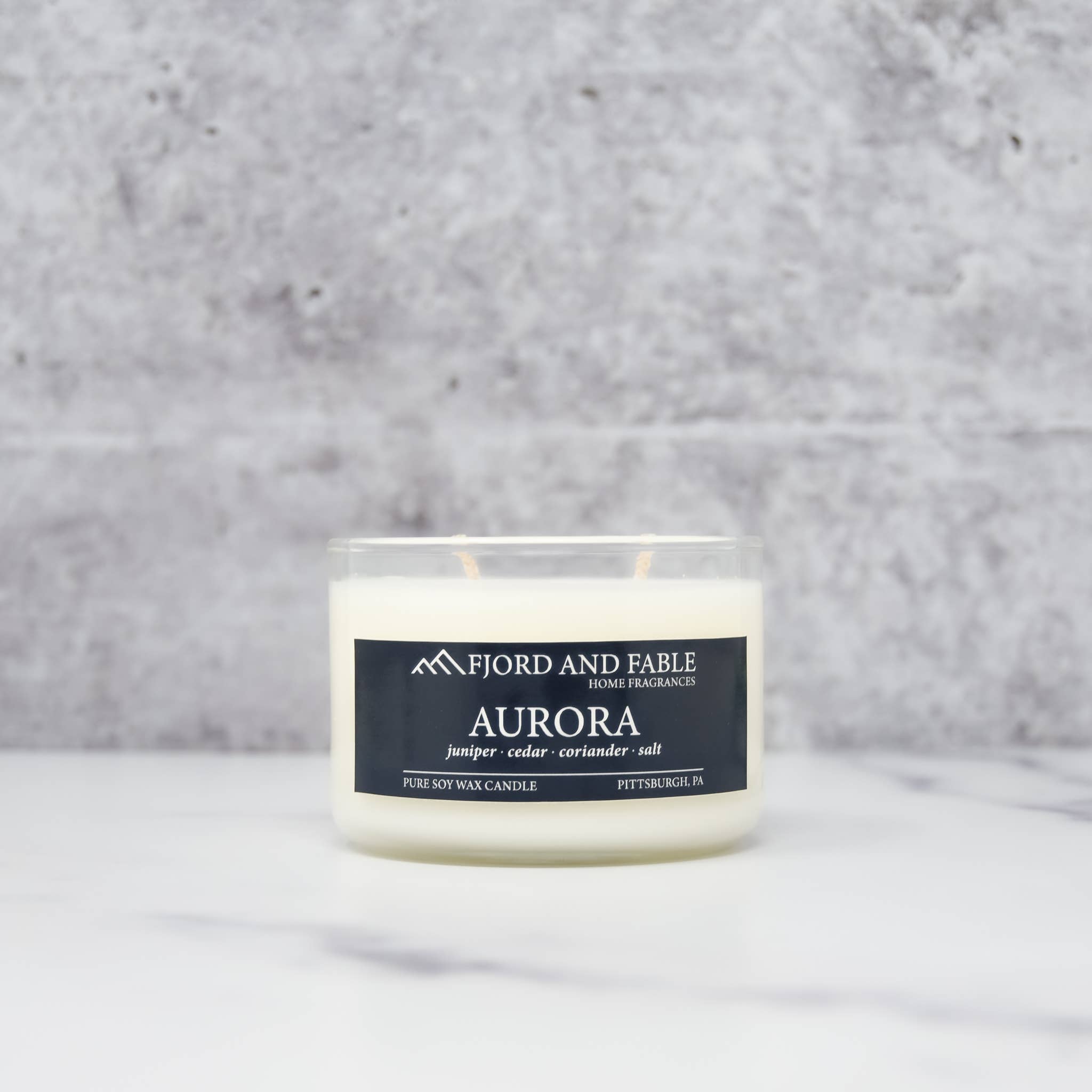 A white 4oz Aurora Premium Soy Candle by Fjord and Fable, featuring a juniper cedar coriander salt scent, sits elegantly on a marble surface against a gray stone background in its sleek glass jar.