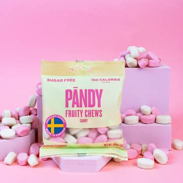 On display is a 1.8oz bag of Pandy Sugar Free Fruity Chews, surrounded by scattered pink and white chewy candies against a pink background. The packaging highlights that they contain only 100 calories and are sugar-free gummies.