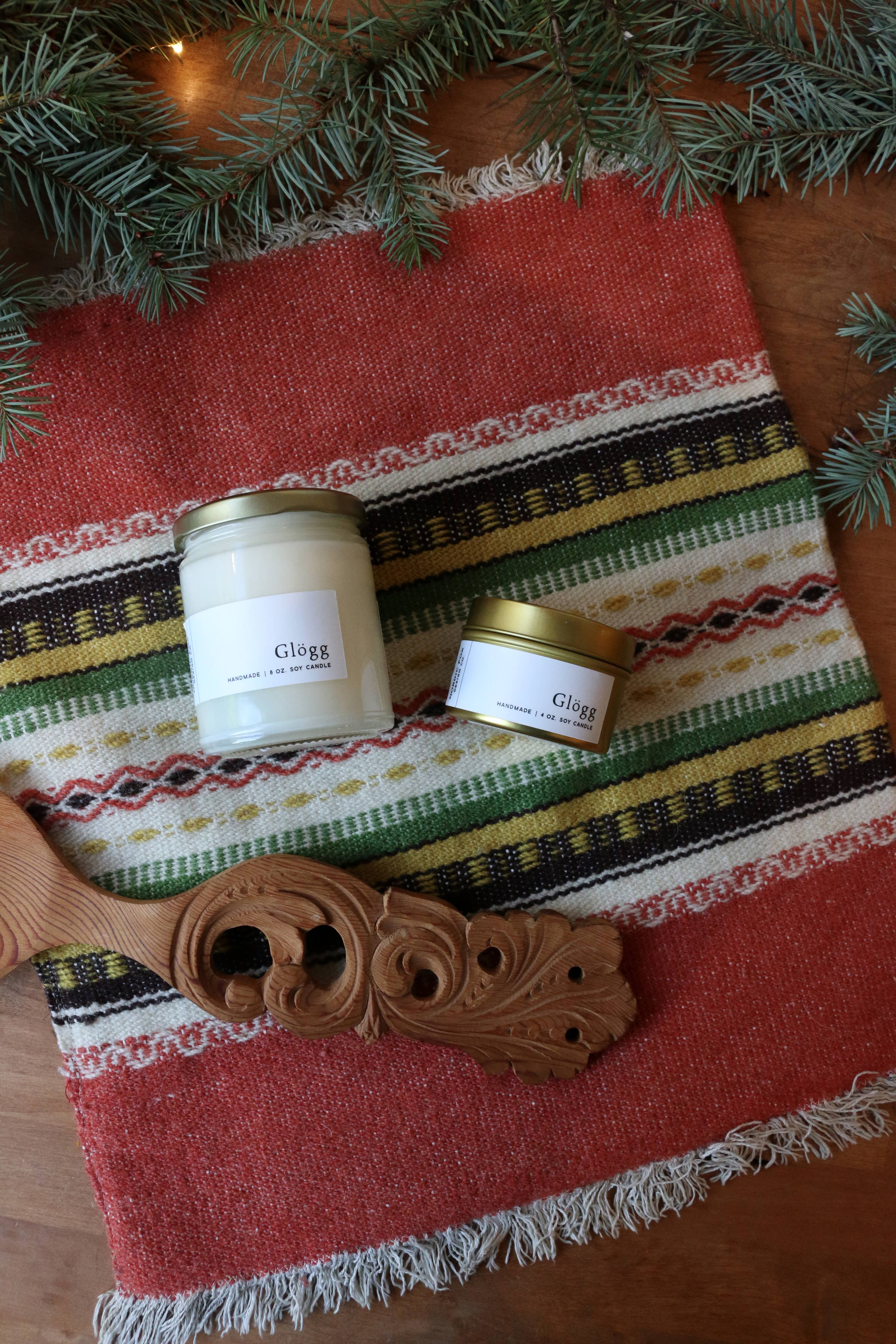 Two 8 oz. Glögg Handmade Soy Candles in glass jars sit on a red and green patterned textile beside a carved wooden object, with pine branches above.