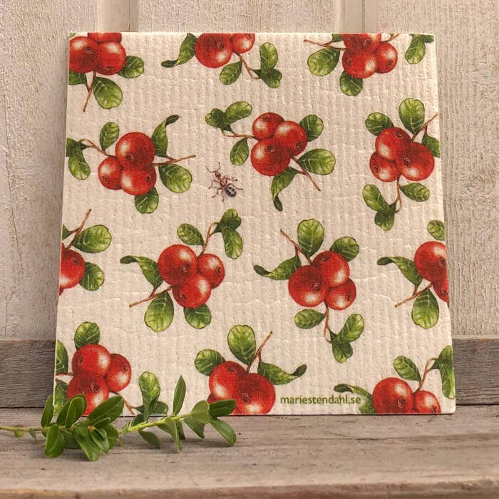 A Lingonberries dishcloth by Marie Stendahl is showcased against a wooden backdrop. Featuring an eco-friendly design, this biodegradable cloth includes a small insect motif, with a green plant placed nearby.