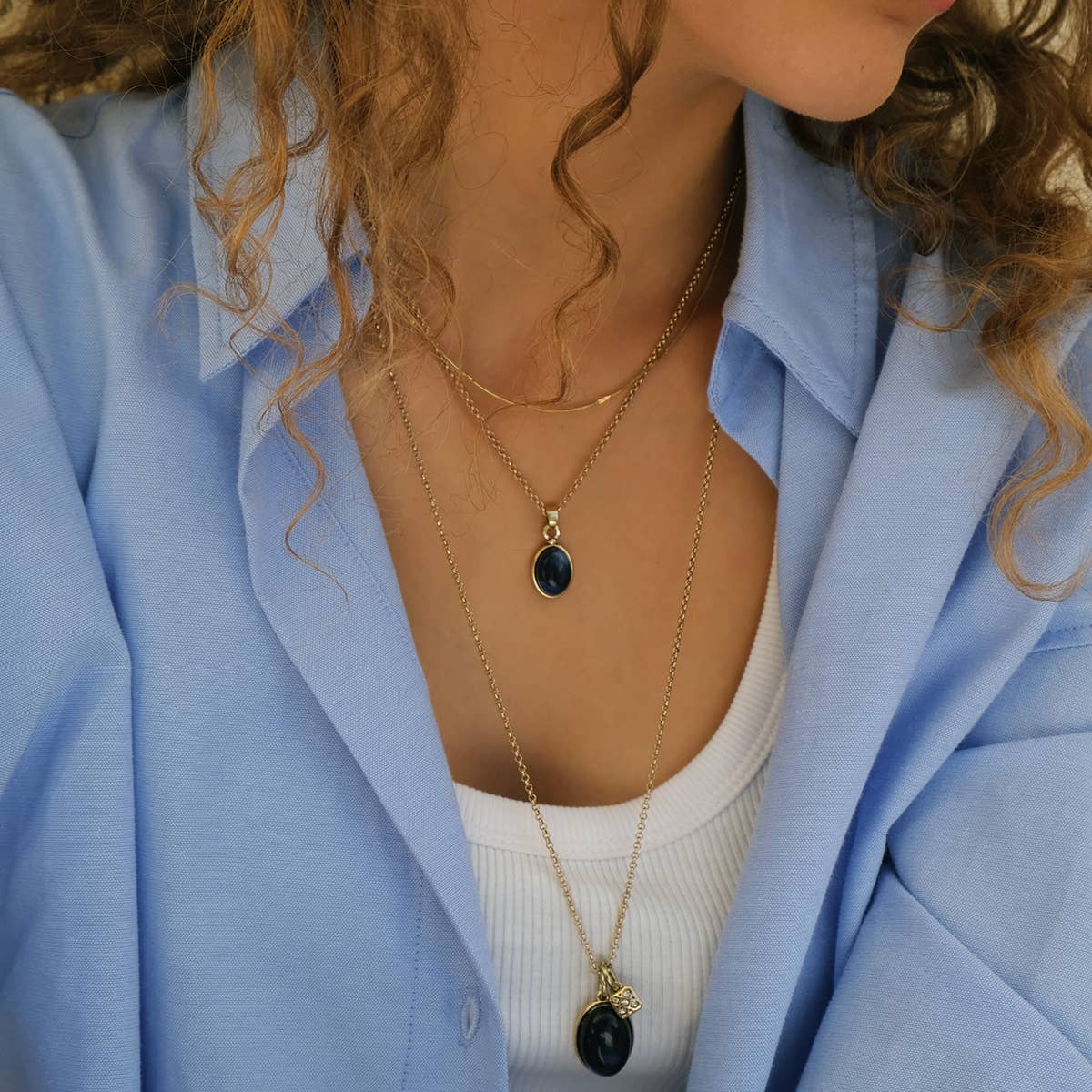 The woman exudes elegance in a blue shirt and white top, enhanced by two A&C Oslo Gold Plated Glass and Crystal Pendant Necklaces. Their stunning crystal pendants add brilliance to her ensemble.