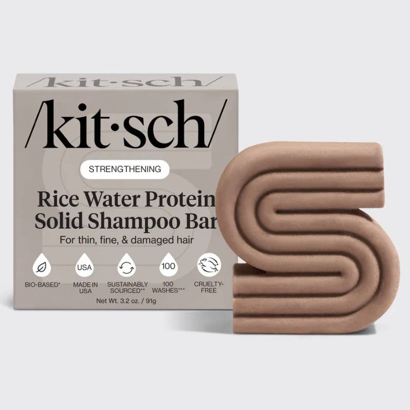 A brown, S-shaped solid shampoo bar is shown in front of a box labeled "KITSCH Shampoo: Rice Water Protein Solid Shampoo Bar for Hair Growth 3.2oz," formulated for thin, fine, and damaged hair. The packaging features various icons highlighting its benefits. This sustainable shampoo bar is crafted to promote hair growth and boost volume while supporting healthy hair and environmental care.