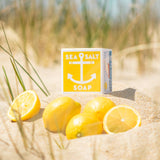 A 4 oz bar of Swedish Dream Sea Salt Lemon Soap featuring a yellow anchor design rests on sand beside whole and sliced lemons, with beach grass in the background.