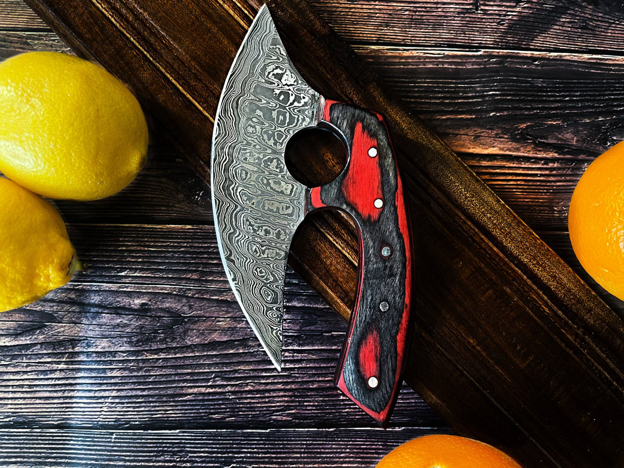 An Alaskan Ulu style Damascus steel knife with a red and black diamond wood handle rests on a wooden surface next to lemons and oranges.