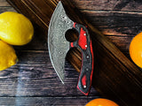 An Alaskan Ulu style Damascus steel knife with a red and black diamond wood handle rests on a wooden surface next to lemons and oranges.