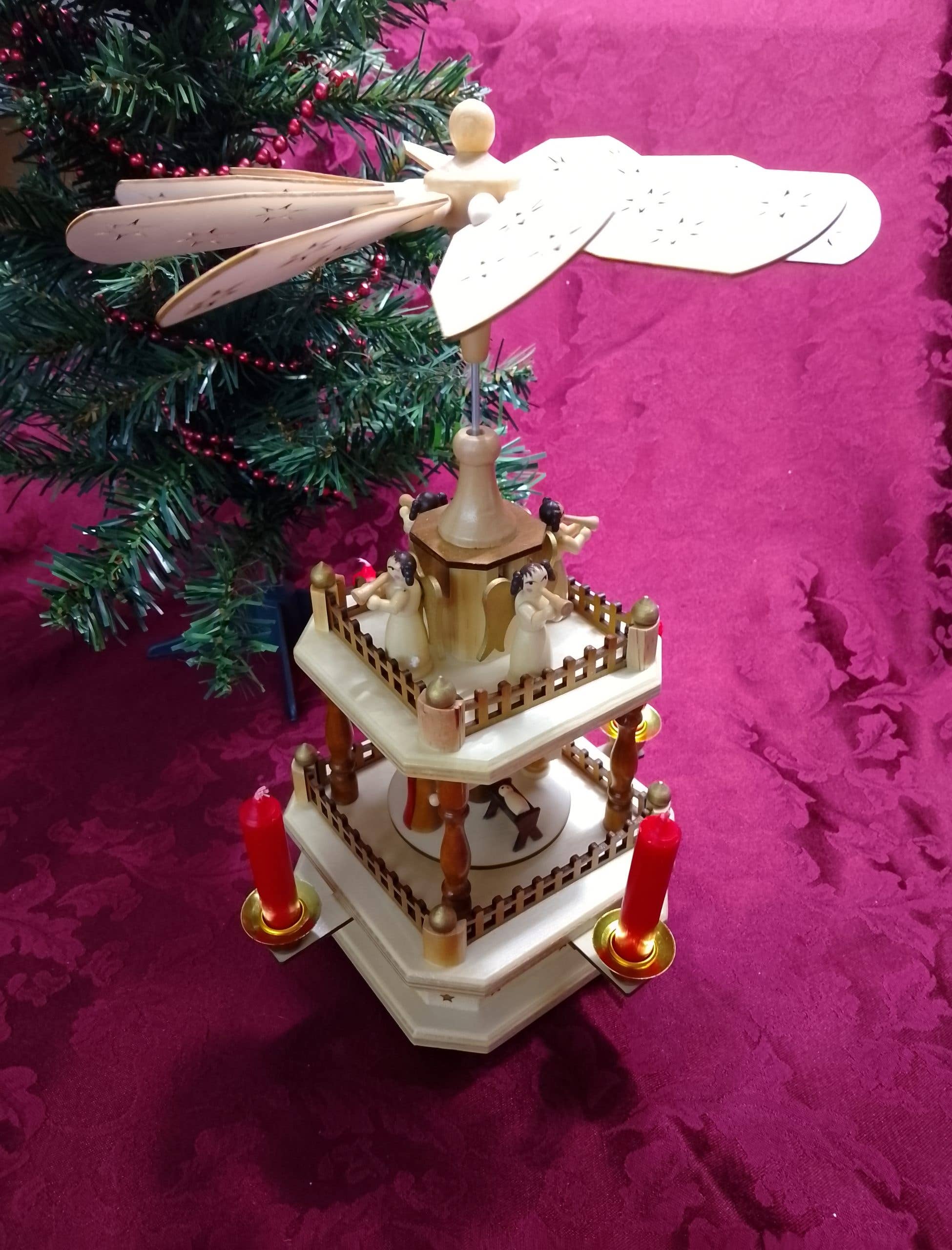 A "Christmas Pyramid: Nativity Square," adorned with candles and small nativity figurines alongside a spinning propeller, rests on a red tablecloth. In the background, a Christmas tree partially emerges, enhancing the festive atmosphere.