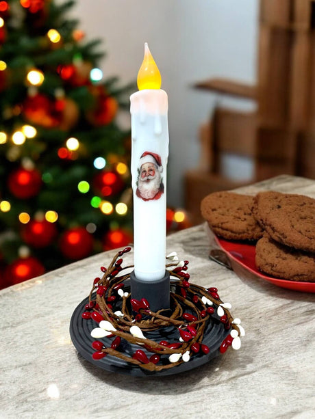 A Santa Christmas LED Battery Operated Timer Taper Candle in white sits by a plate of cookies, softly glowing on the table with the Christmas tree twinkling in the background.