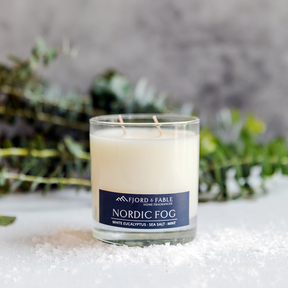 An 8oz Nordic Fog Candle sits on a white surface with scattered salt and green foliage, evoking the fresh scent of eucalyptus mint in the background.