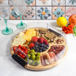 Round bamboo charcuterie board with a clear plastic lid, divided into sections containing cheese, berries, nuts, olives, tomatoes, and meat slices.