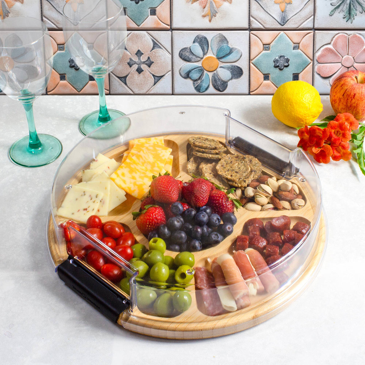Round bamboo charcuterie board with a clear plastic lid, divided into sections containing cheese, berries, nuts, olives, tomatoes, and meat slices.