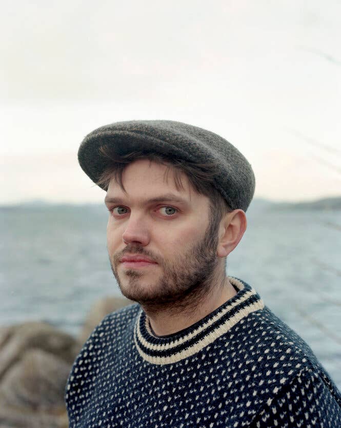 A person wearing a men's blue wool flat cap and patterned sweater stands near a body of water with rocks in the background.