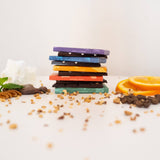 Stack of assorted chocolate bars with different colors, surrounded by nuts, chocolate pieces, coffee beans, and orange slices on a white background.