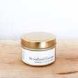 A small, gold tin of Candle: Woodland Gnome Handmade Soy Candle (4 oz.) sits on a light wooden surface, its earthy scent and natural soy wax highlighted against a plain white background.